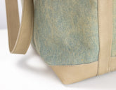 Close-up of a tote bag corner showing a textured sage green canvas body with tan leather trim and strap on a white background.
