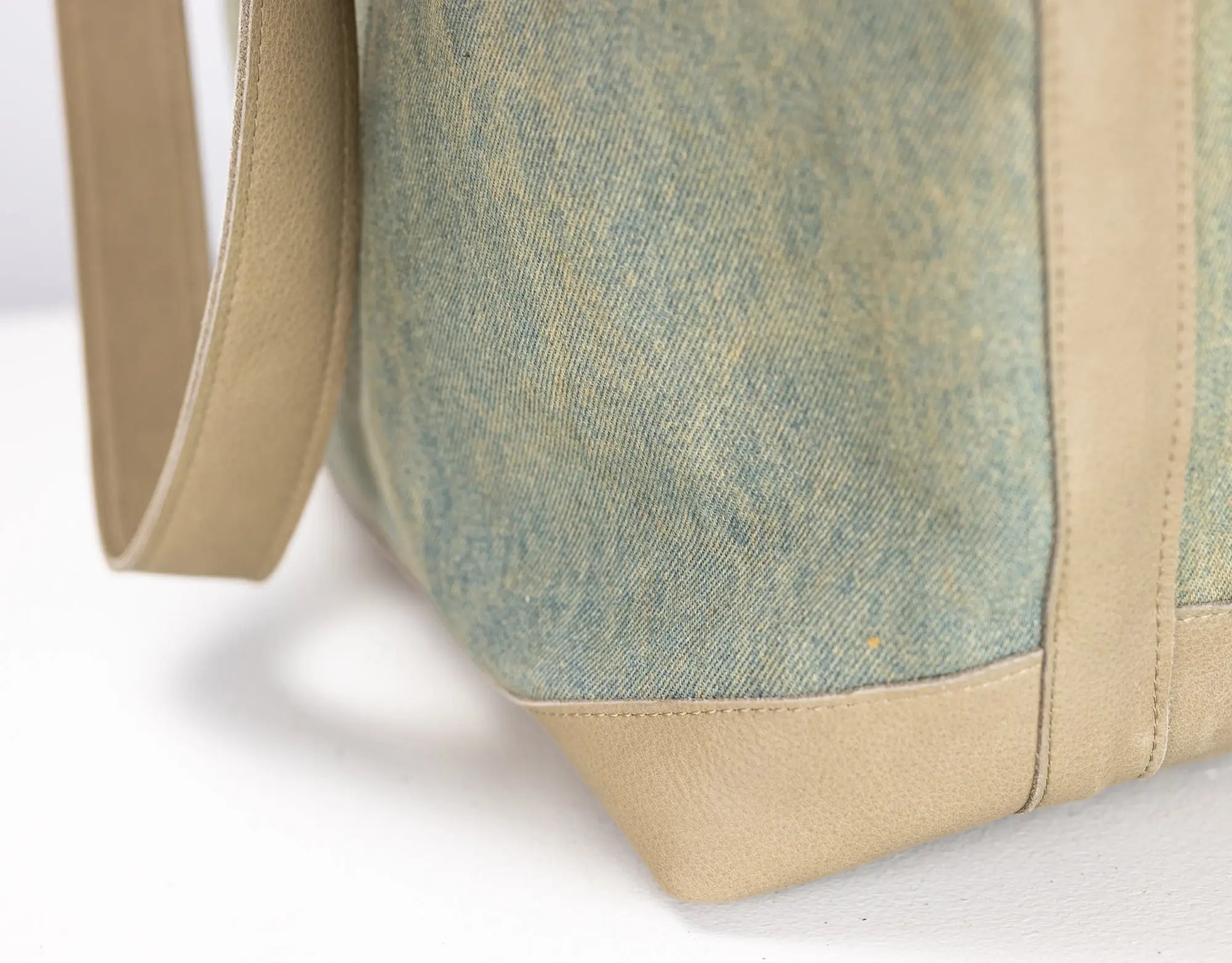 Close-up of a tote bag corner showing a textured sage green canvas body with tan leather trim and strap on a white background.