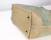 Close-up of a handbag corner showing a beige leather base with two brass rivets and a light blue-green woven fabric side panel on a white background.