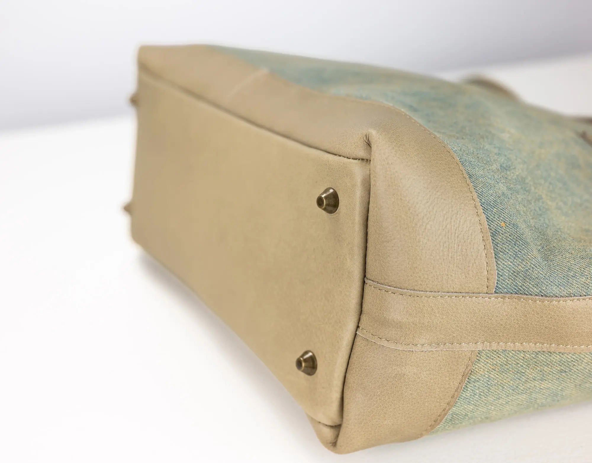 Close-up of a handbag corner showing a beige leather base with two brass rivets and a light blue-green woven fabric side panel on a white background.