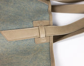 Close-up of a tote bag showing a textured blue-gray fabric body with tan leather trim and strap details on a white background.