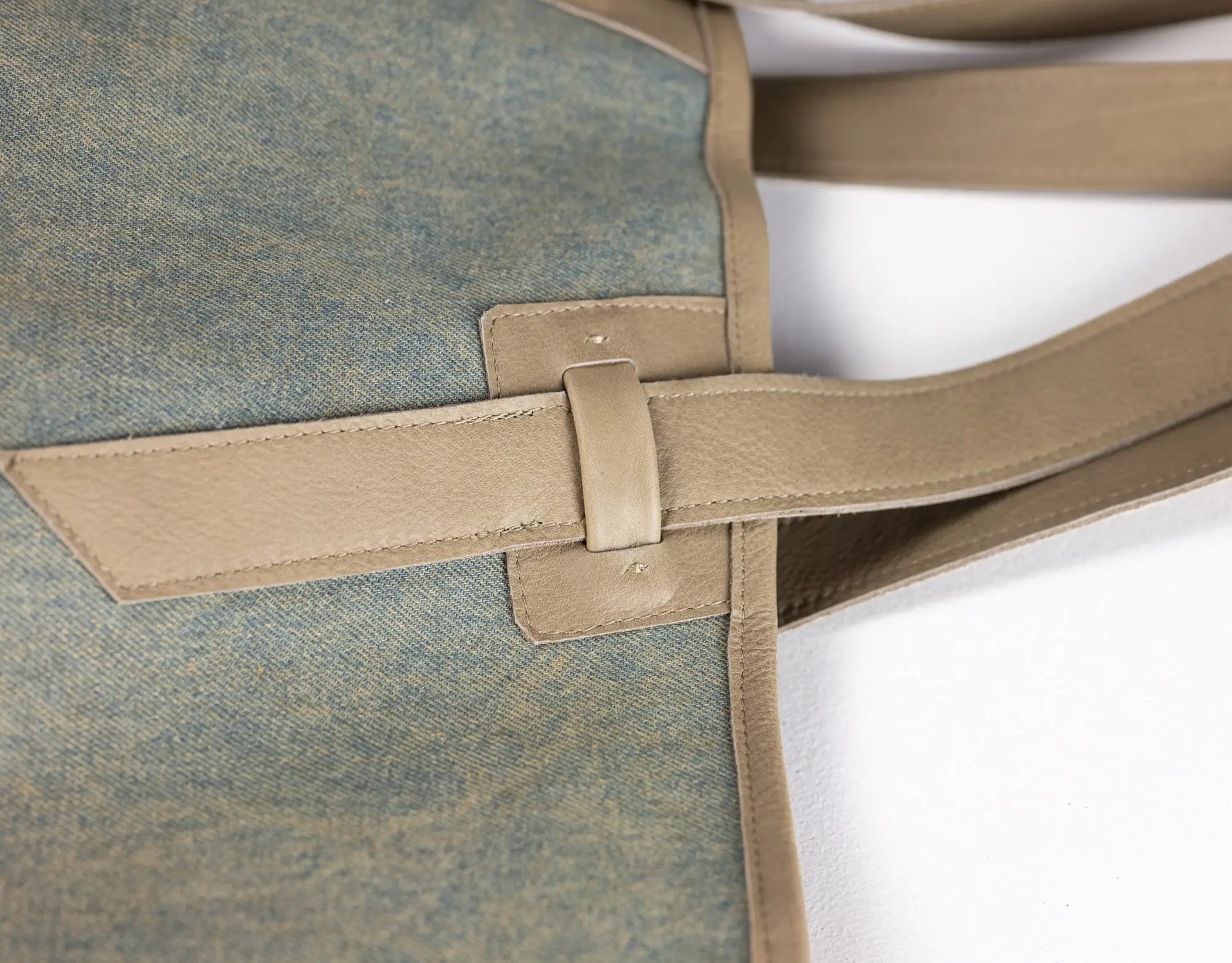 Close-up of a tote bag showing a textured blue-gray fabric body with tan leather trim and strap details on a white background.