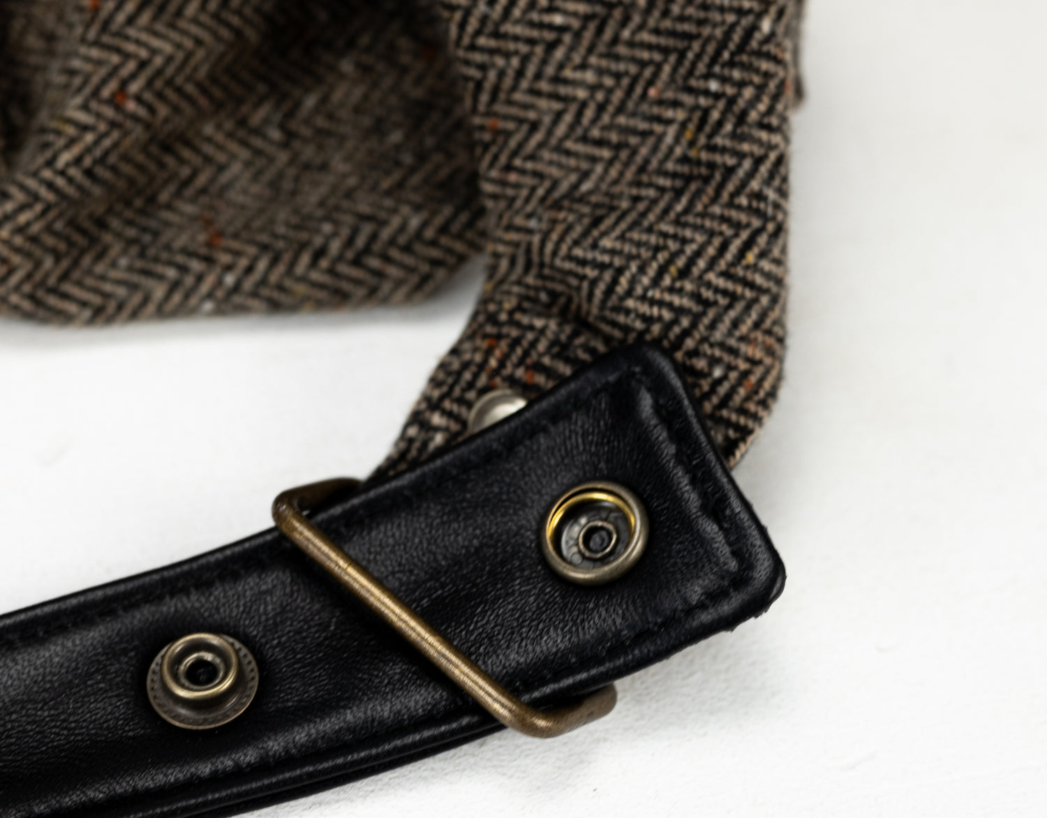 Close-up of a herringbone-patterned fabric with a black leather strap and brass buckle on a white background.