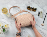 Person holding a woven beige handbag on a marble surface with a glass of wine, sunglasses, and other items.
