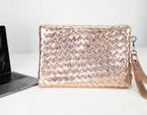 Rose gold woven clutch bag on a white surface with a laptop.