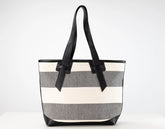 A striped tote bag with horizontal bands of cream, medium gray, and white, featuring black leather handles and trim, set against a plain white background.