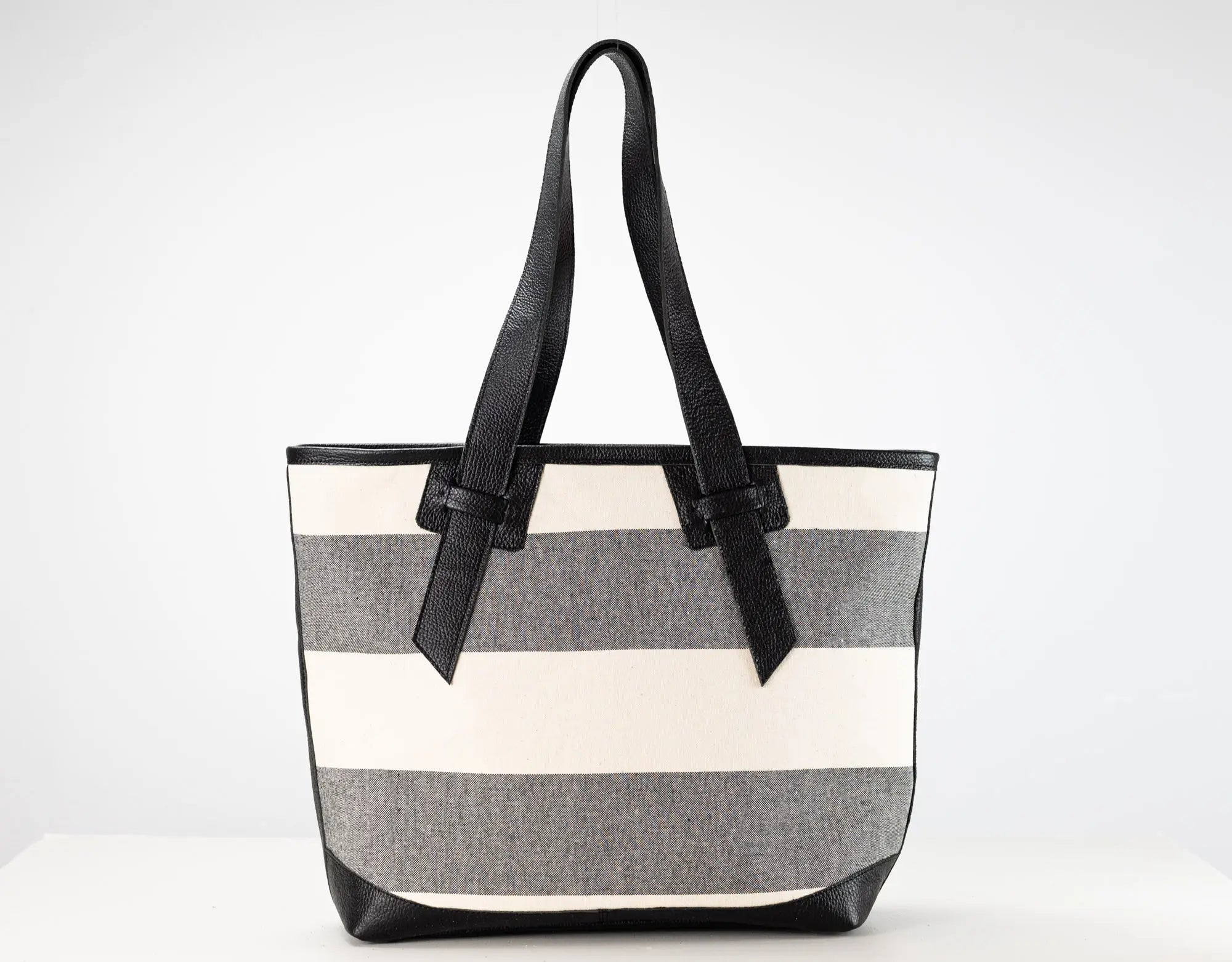 A striped tote bag with horizontal bands of cream, medium gray, and white, featuring black leather handles and trim, set against a plain white background.