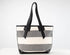 A striped tote bag with horizontal bands of cream, medium gray, and white, featuring black leather handles and trim, set against a plain white background.