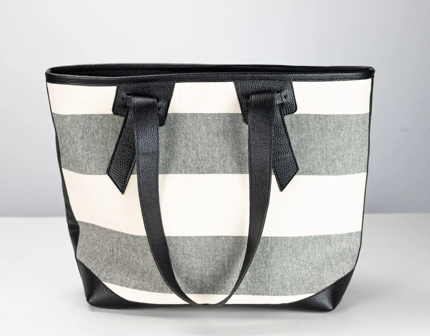 A striped tote bag with horizontal gray and off-white stripes, featuring black leather handles and trim, set against a neutral light gray background.