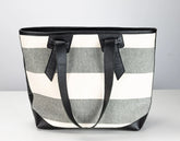 A striped tote bag with horizontal gray and off-white stripes, featuring black leather handles and trim, set against a neutral light gray background.
