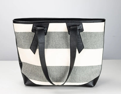 A striped tote bag with horizontal gray and off-white stripes, featuring black leather handles and trim, set against a neutral light gray background.