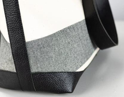Close-up of a structured handbag corner showing black pebbled leather trim and strap against a white and gray woven fabric body.