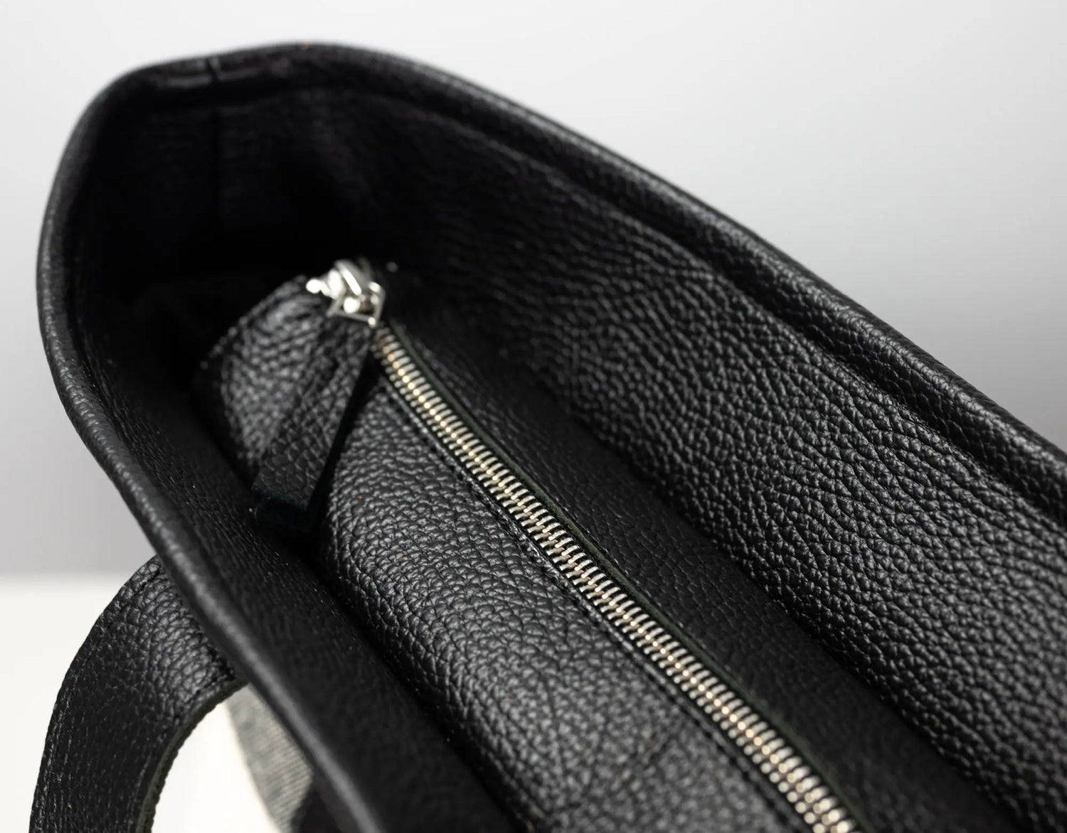 Close-up of the interior of a black pebbled leather bag, showing a silver zipper and a small matching leather pull tab against a light gray background.