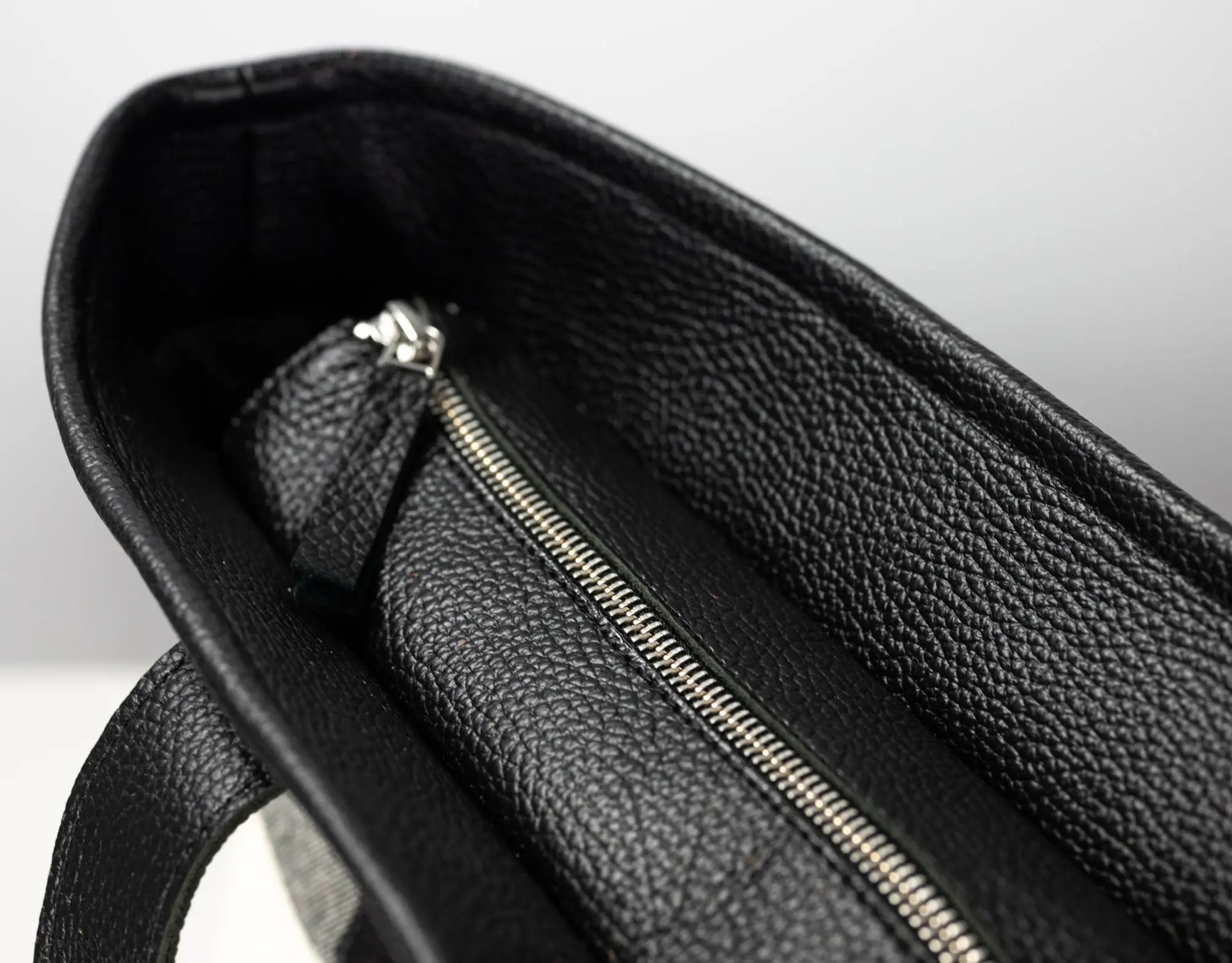 Close-up of the interior of a black pebbled leather bag, showing a silver zipper and a small matching leather pull tab against a light gray background.