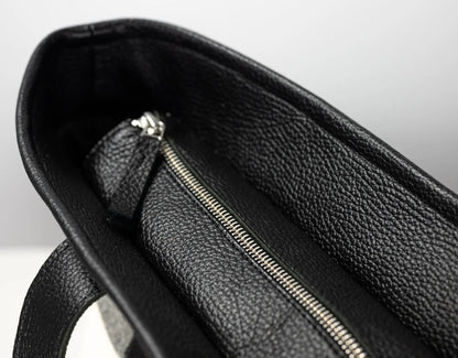 Close-up of the interior of a black pebbled leather bag, showing a silver zipper and a small matching leather pull tab against a light gray background.