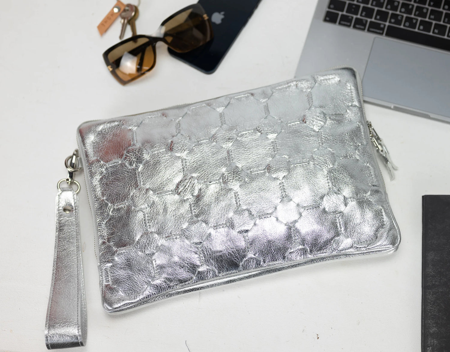 Silver clutch bag on a white surface with sunglasses, phone, and laptop in the background