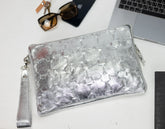 Silver clutch bag on a white surface with sunglasses, phone, and laptop in the background