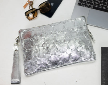 Silver clutch bag on a white surface with sunglasses, phone, and laptop in the background
