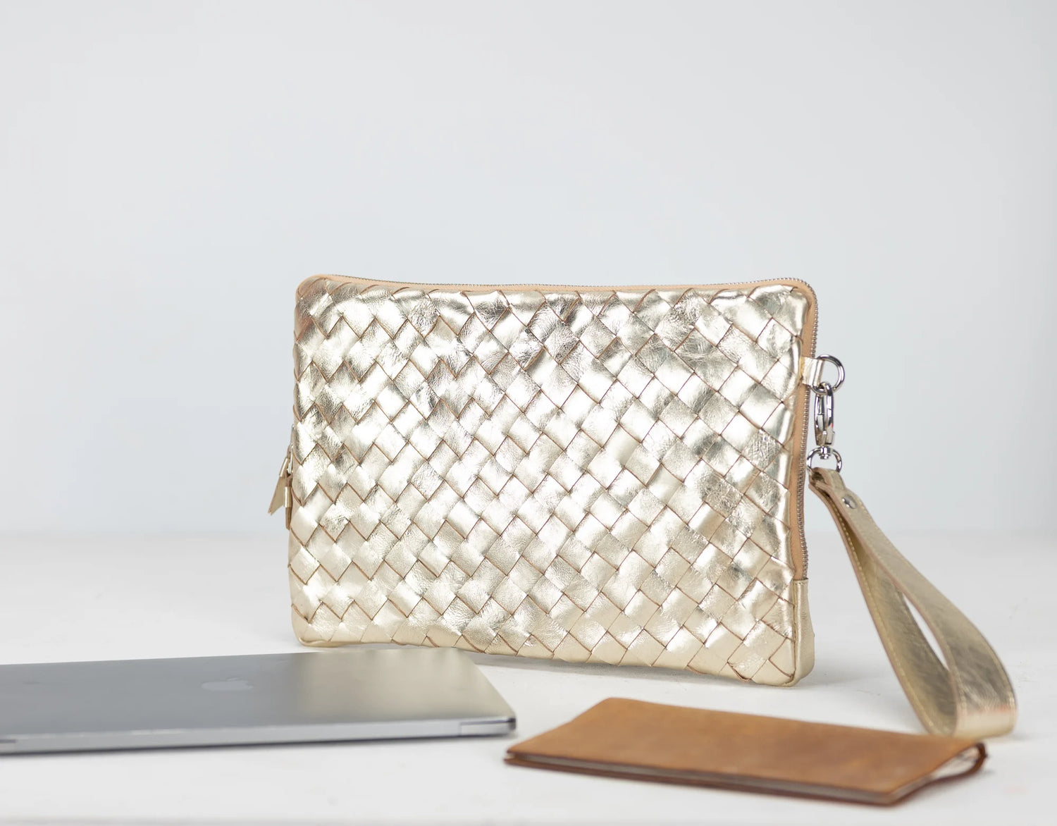Gold woven clutch bag with a silver laptop and brown wallet on a light gray background