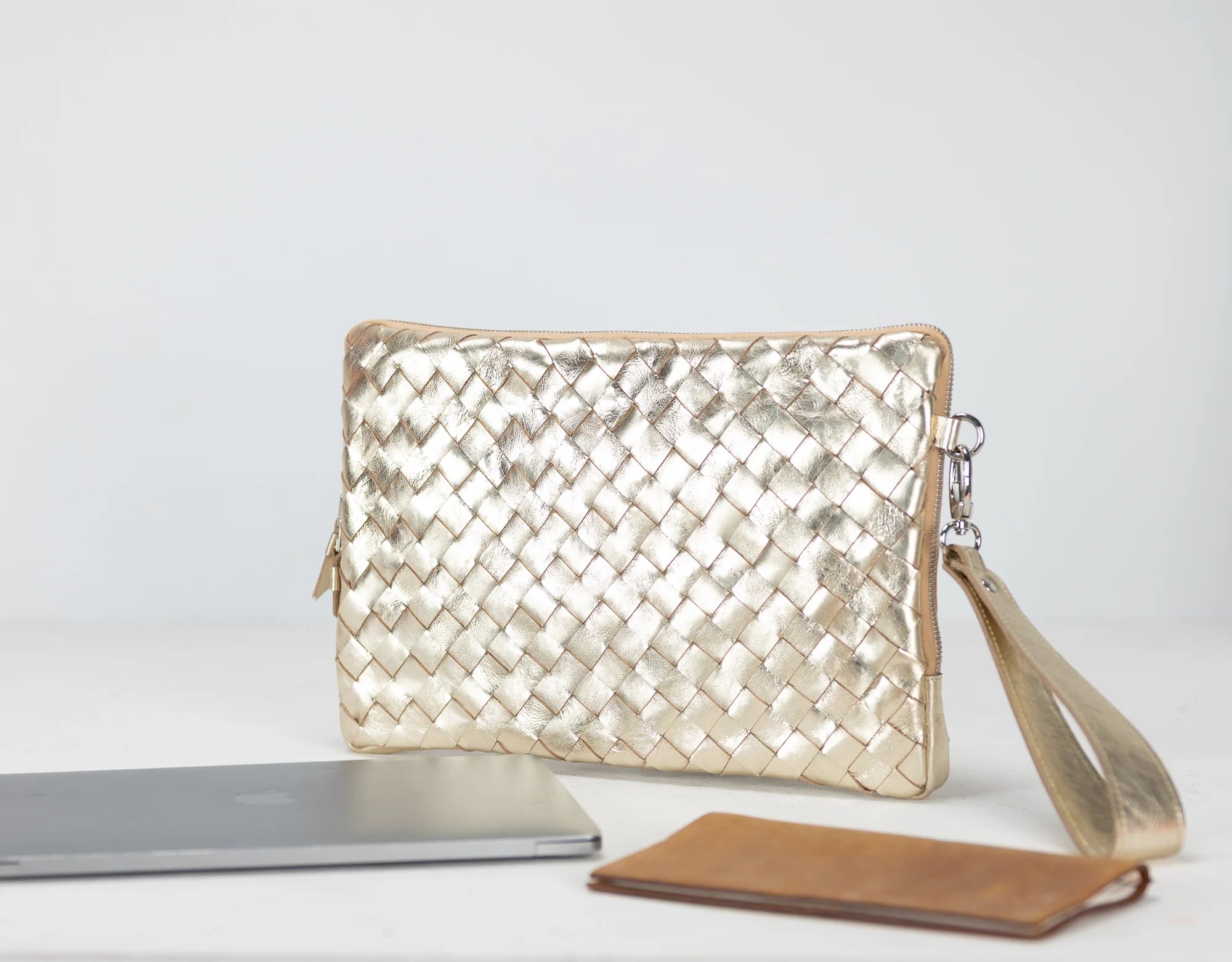 Gold woven clutch bag with a silver laptop and brown wallet on a light gray background