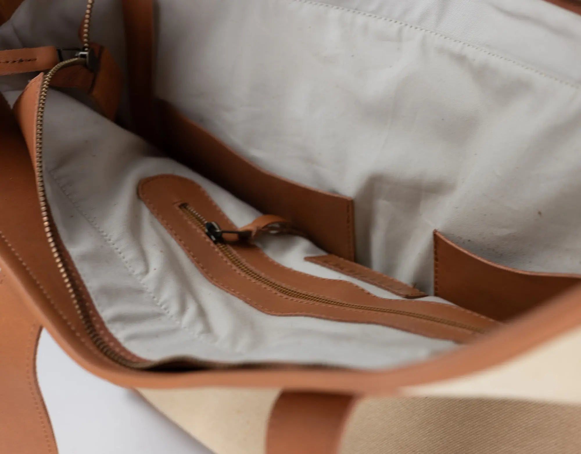 Close-up of the interior of a tote bag showing a light beige fabric lining, tan leather trim, and a zippered pocket.