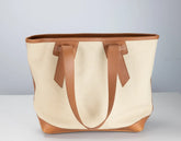A canvas tote bag with natural cream fabric and tan leather trim, handles, and base, photographed against a neutral light gray background.