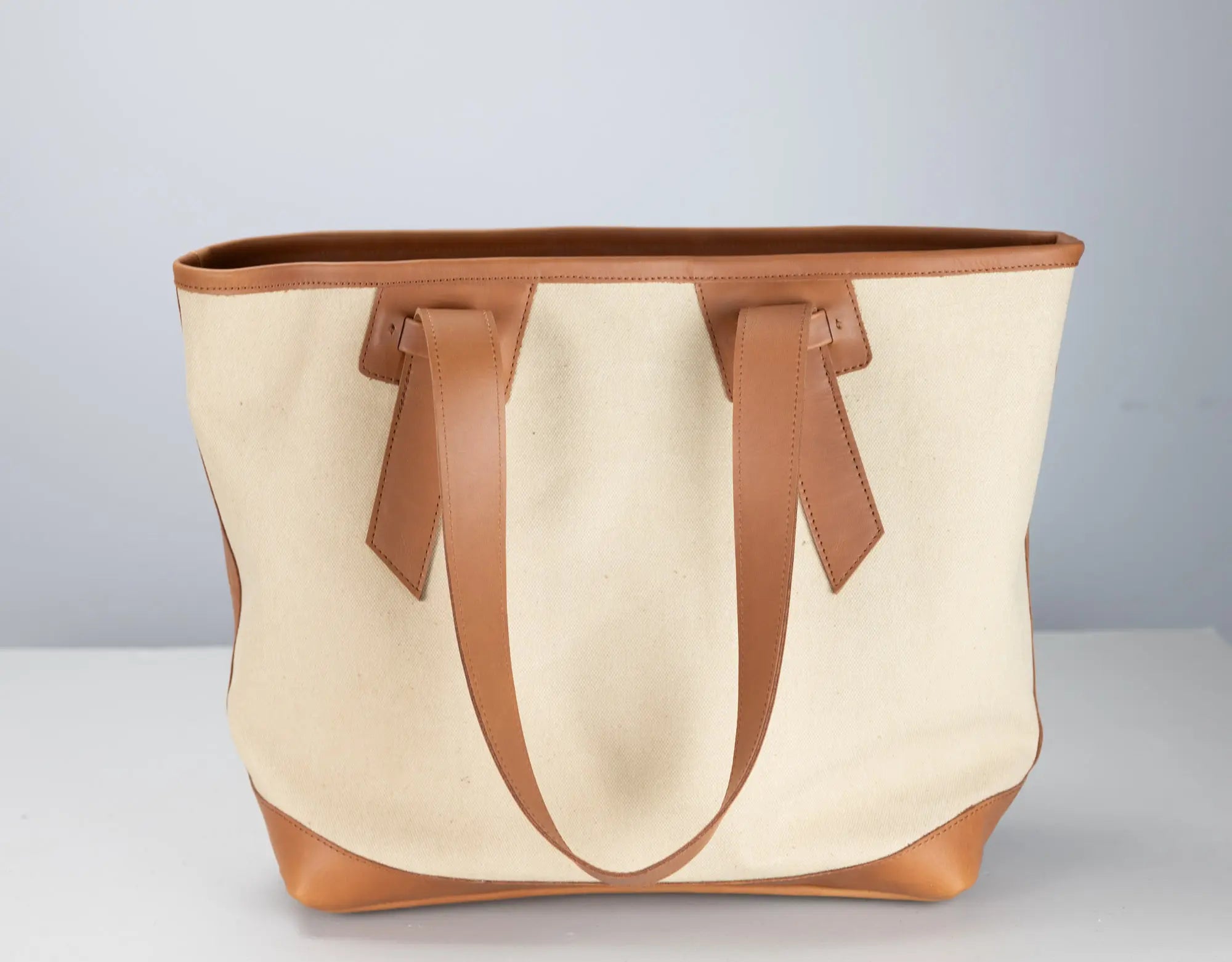 A canvas tote bag with natural cream fabric and tan leather trim, handles, and base, photographed against a neutral light gray background.