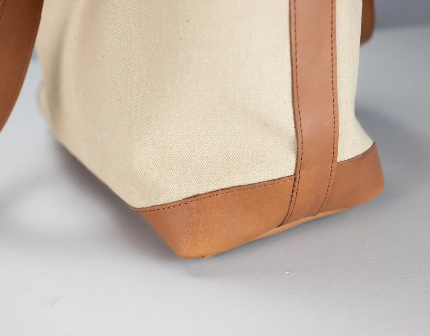 Close-up of a canvas tote bag corner showing a natural beige fabric body with tan leather trim and a matching leather handle on a light gray background.