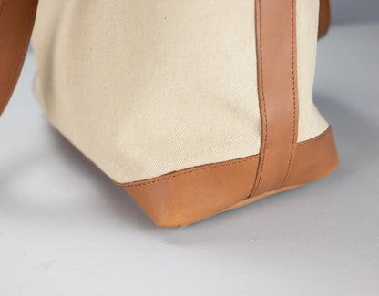 Close-up of a canvas tote bag corner showing a natural beige fabric body with tan leather trim and a matching leather handle on a light gray background.