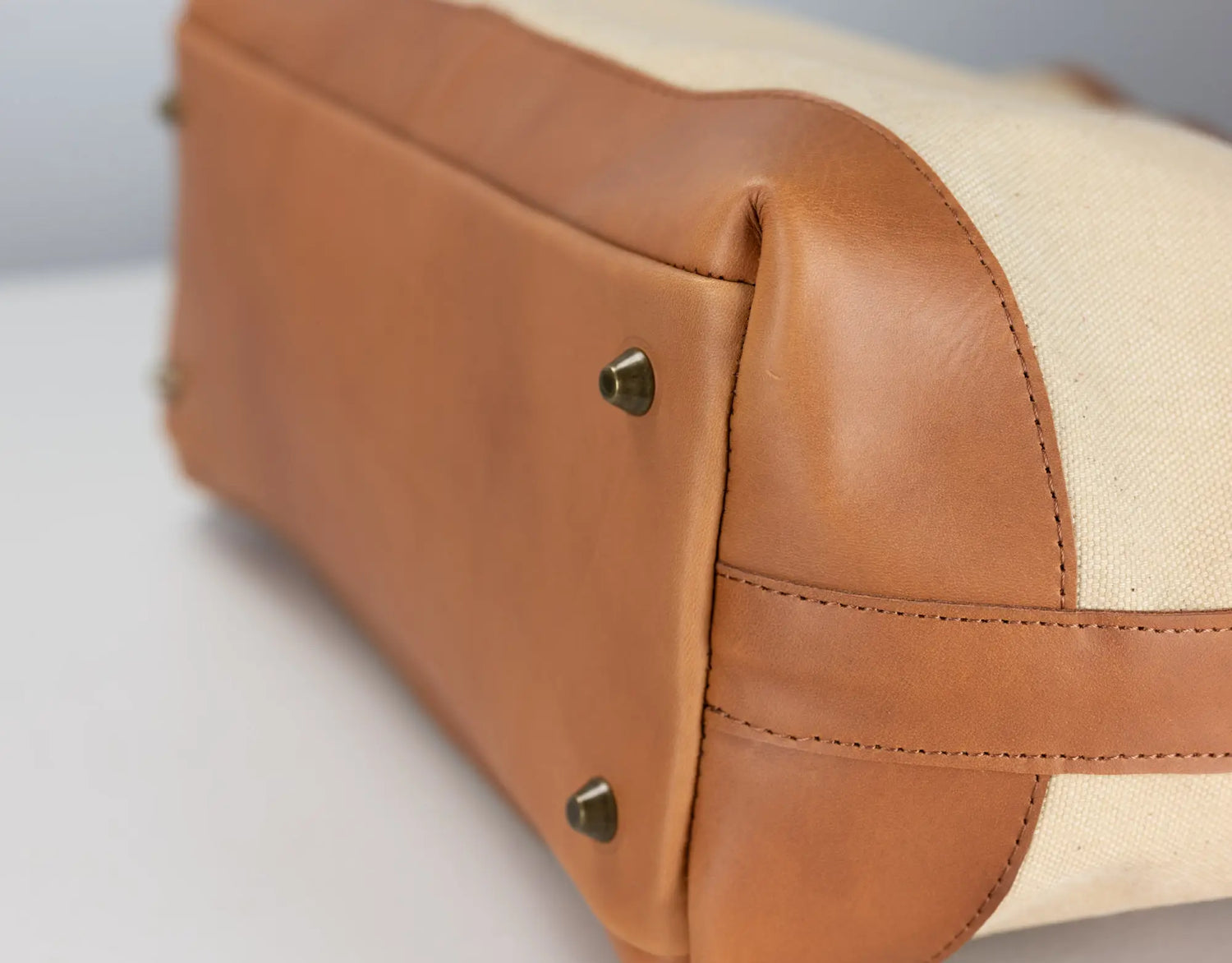 Close-up of a handbag corner showing tan leather trim and a cream canvas body with small metal studs.