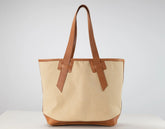 A canvas tote bag with a natural beige body and tan leather handles, trim, and base, photographed against a neutral light gray background.