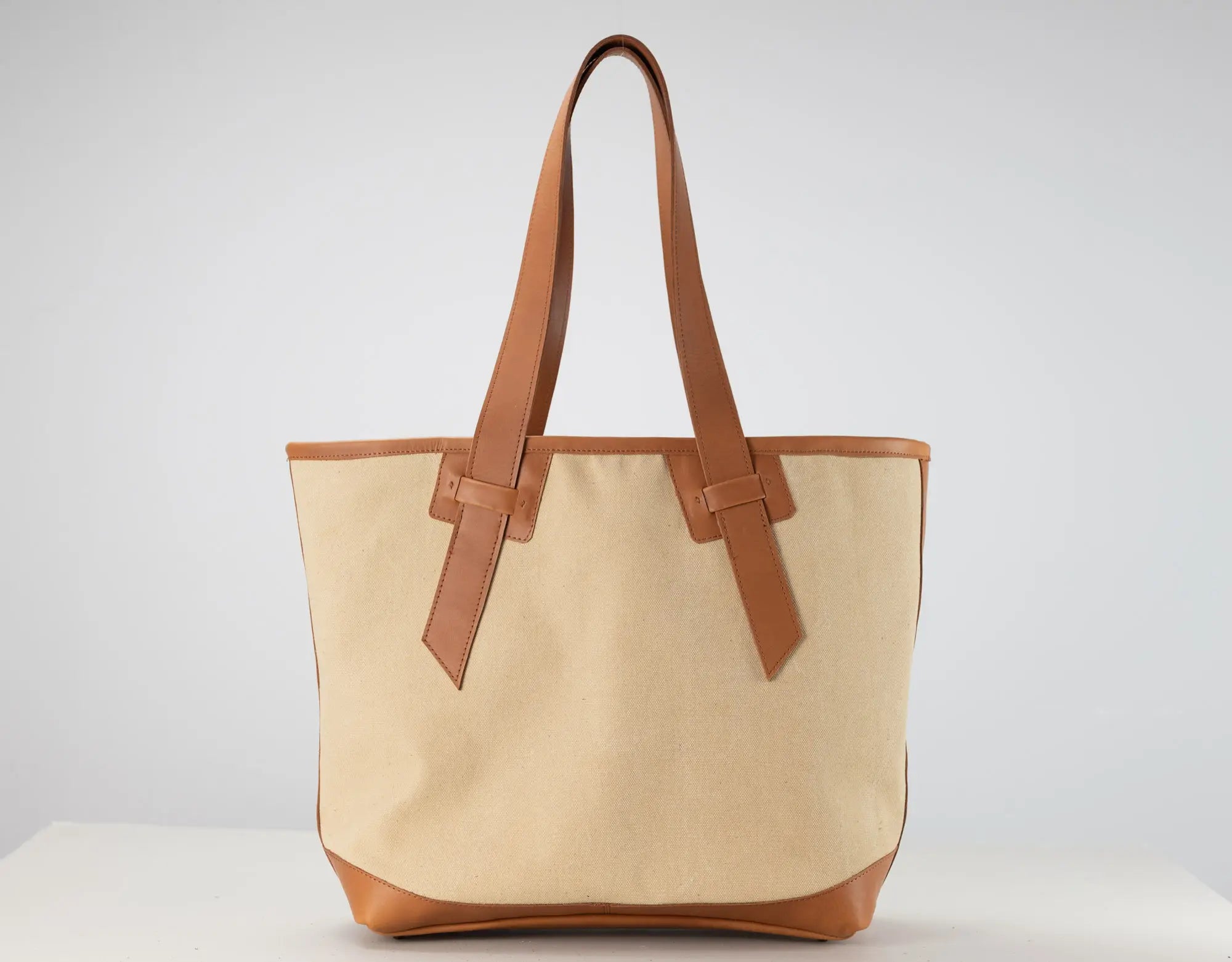 A canvas tote bag with a natural beige body and tan leather handles, trim, and base, photographed against a neutral light gray background.