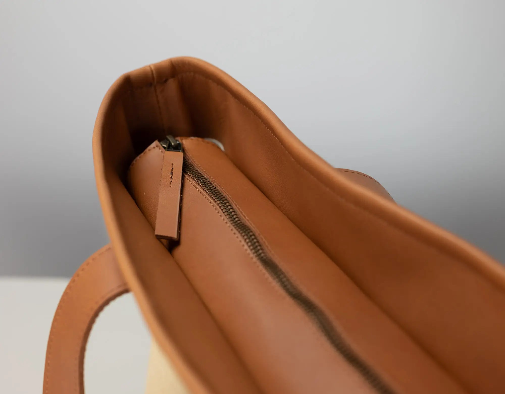 Close-up of the interior of a tan leather tote bag, showing a zippered compartment with a matching leather pull tab against a neutral gray background.