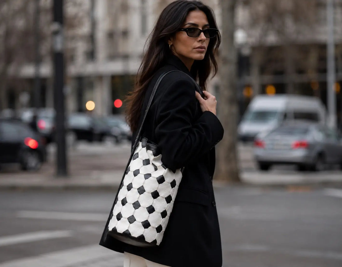 A woman wearing a black coat and sunglasses carries a large black and white checkered leather tote bag on a city street.