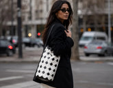 A woman wearing a black coat and sunglasses carries a large black and white checkered leather tote bag on a city street.