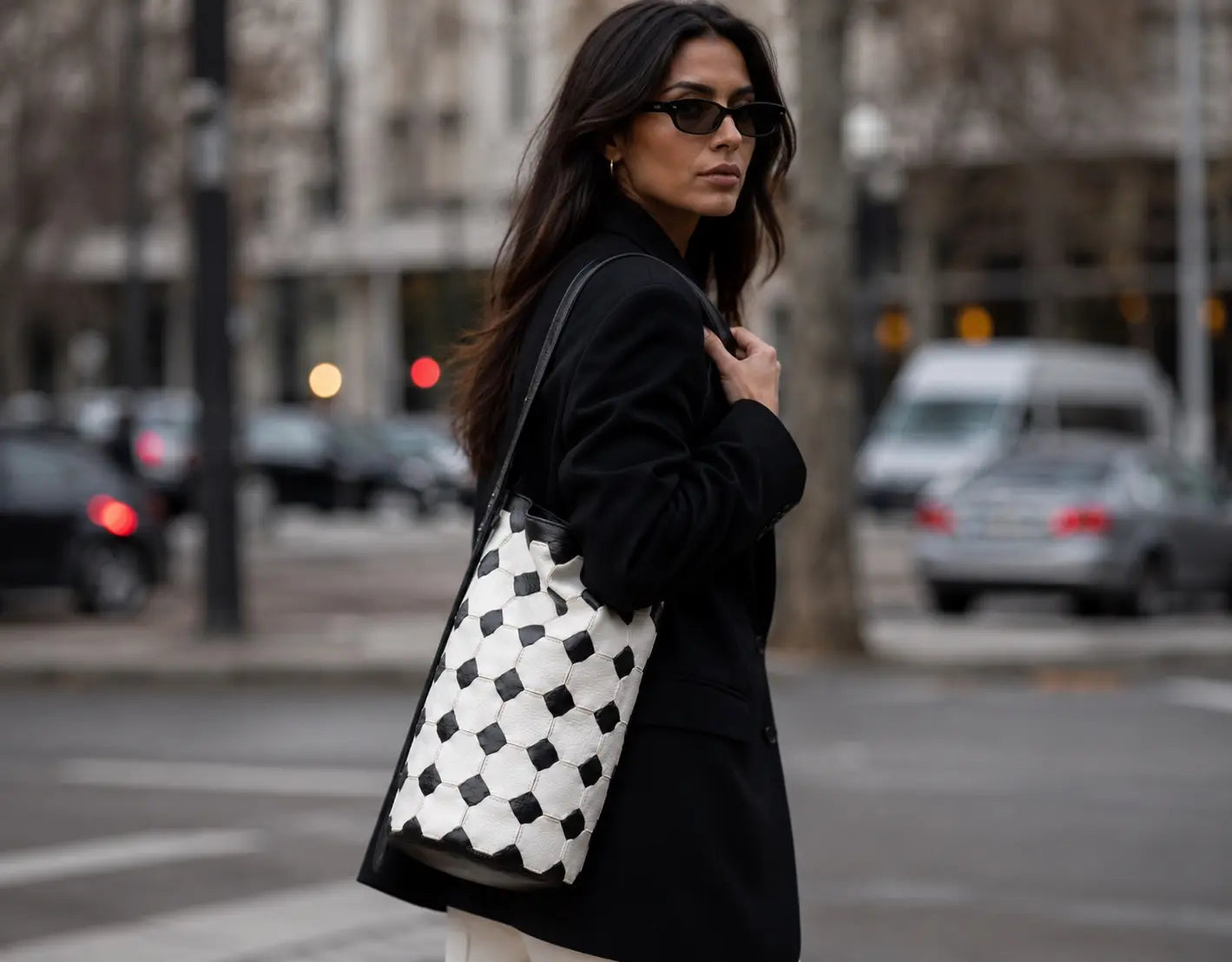 A woman wearing a black coat and sunglasses carries a large black and white checkered leather tote bag on a city street.