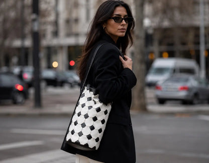 A woman wearing a black coat and sunglasses carries a large black and white checkered leather tote bag on a city street.