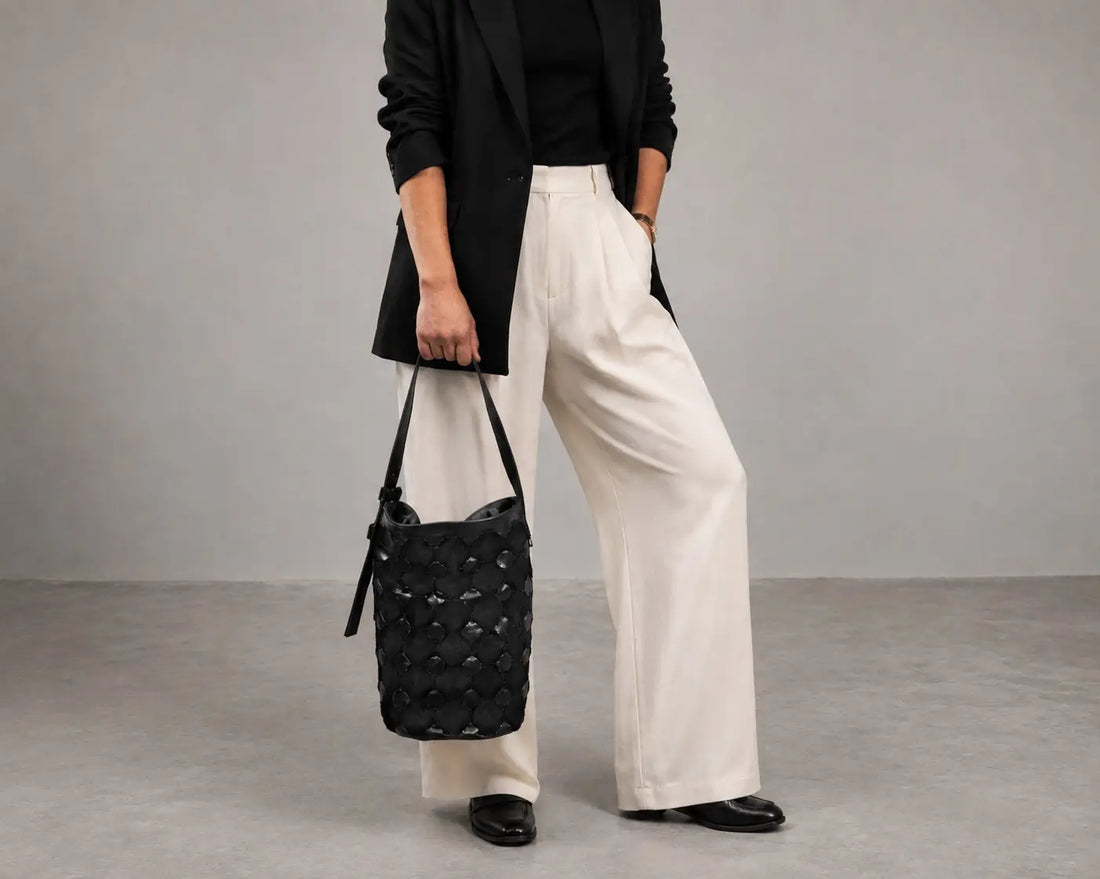 A model wearing a black blazer, black top, and wide-leg cream trousers holds a black quilted bucket bag with a shoulder strap against a neutral gray studio background.