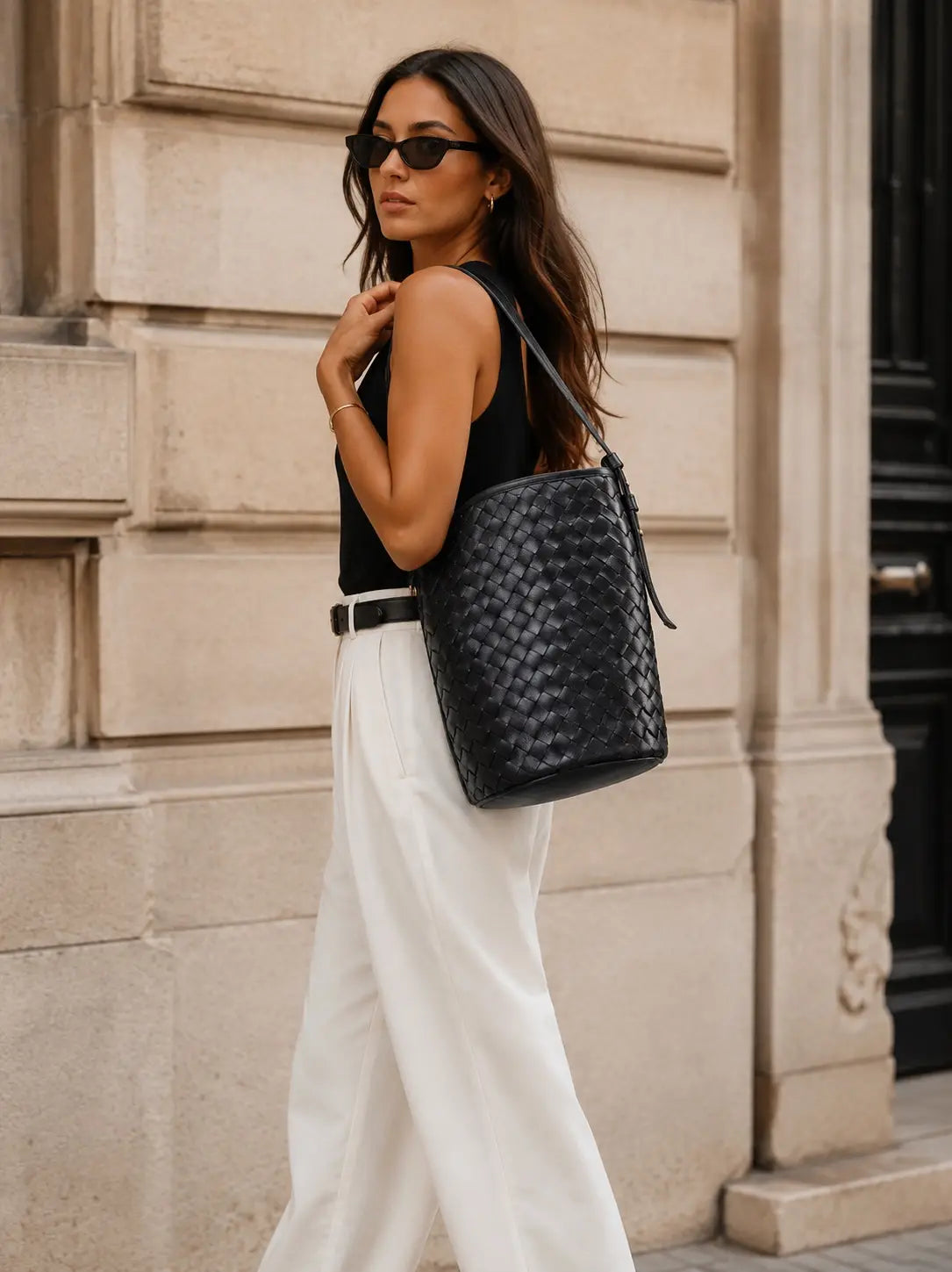 A woman wearing a black sleeveless top, white wide-leg trousers, and black sunglasses carries a black woven leather shoulder bag with a thin black strap, walking past a beige stone building.