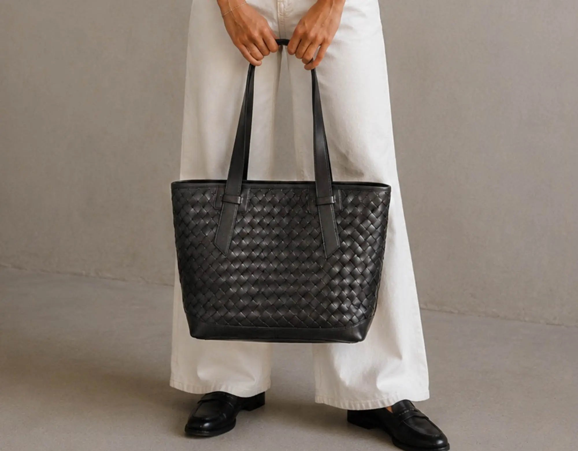 A person wearing white wide-leg trousers and black loafers holds a large black woven leather tote bag against a neutral gray studio background.