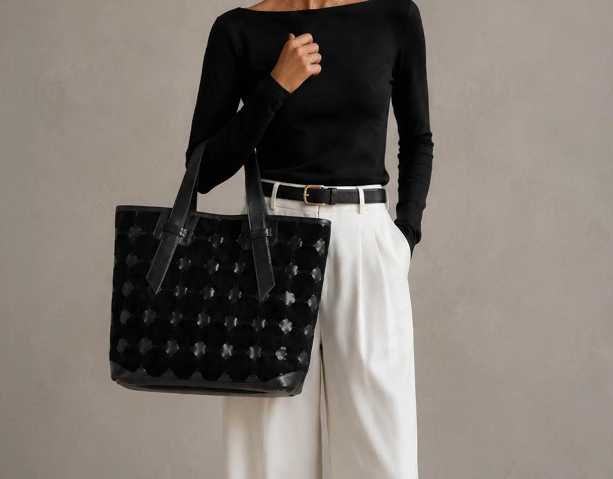 A model wearing a black long-sleeve top and white wide-leg trousers carries a large black woven tote bag with a scalloped edge and black shoulder straps against a neutral gray background.