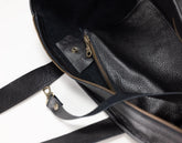 Close-up of a black pebbled leather bag interior showing a zippered pocket with brass-toned hardware and a matching black leather strap on a white background.