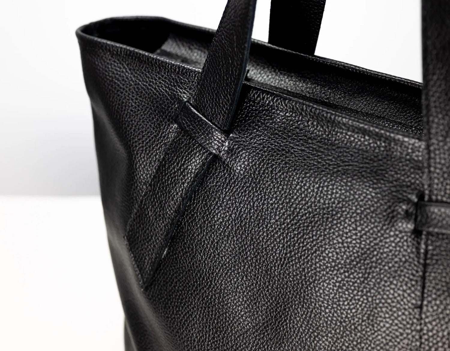 Close-up of a black pebbled leather tote bag showing the texture and strap attachment against a plain white background.