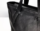 Close-up of a black pebbled leather tote bag showing the texture and strap attachment against a plain white background.