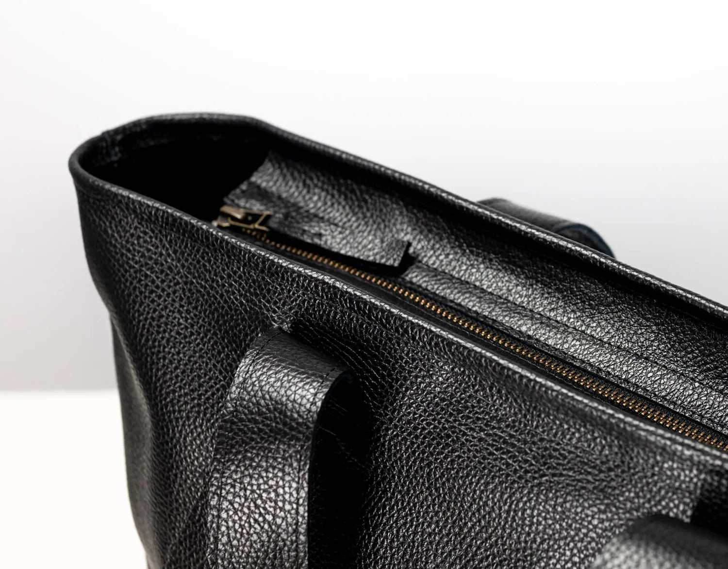 Close-up of a black pebbled leather bag showing the interior opening with a gold-toned zipper and a handle attachment, photographed against a clean white background.