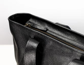 Close-up of a black pebbled leather bag showing the interior opening with a gold-toned zipper and a handle attachment, photographed against a clean white background.