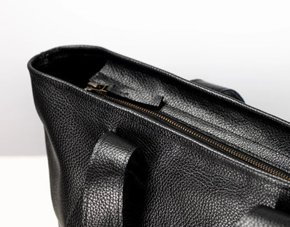 Close-up of a black pebbled leather bag showing the interior opening with a gold-toned zipper and a handle attachment, photographed against a clean white background.