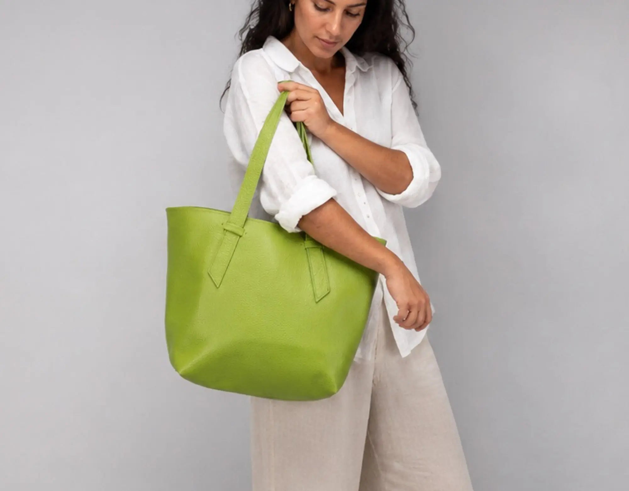 A woman wearing a white button-up shirt and beige trousers holds a large lime green leather tote bag against a plain gray studio background.