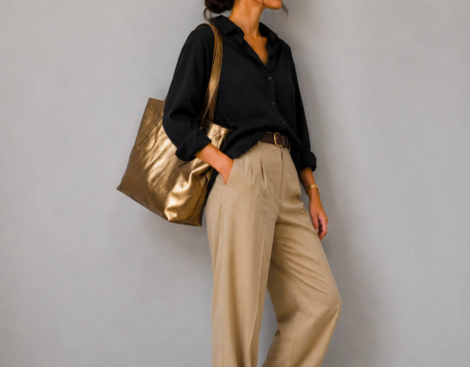 A model wearing a black long-sleeve button-up shirt, beige wide-leg trousers, and carrying a large metallic gold shoulder bag against a plain gray wall.