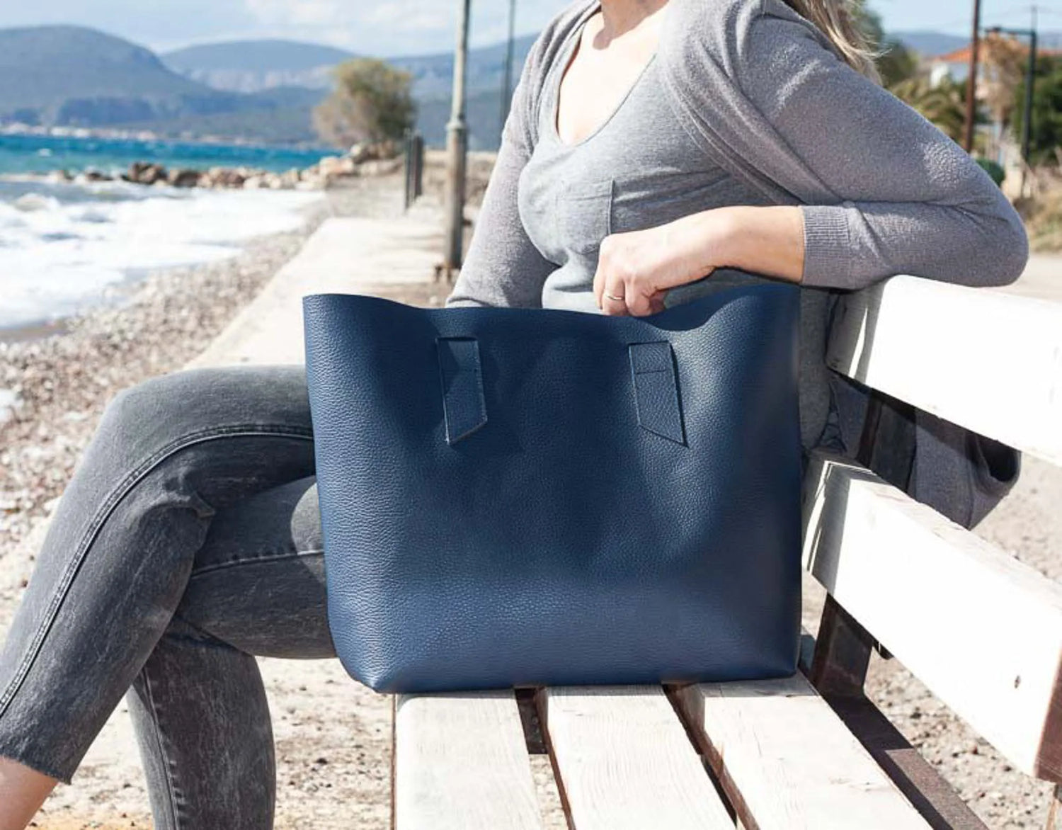 Person sitting on a bench by the sea holding a blue tote bag